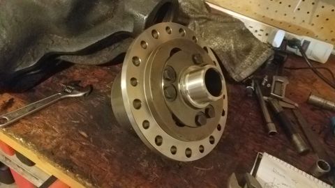 1985 Ford Bronco - A close-up view of a differential part with multiple bolts and a silver-colored flange is shown resting on a wooden workbench. Tools, inclu…