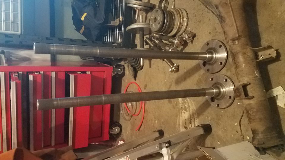 Two steel axle shafts with flanged ends on a concrete floor with a red tool chest and weights.