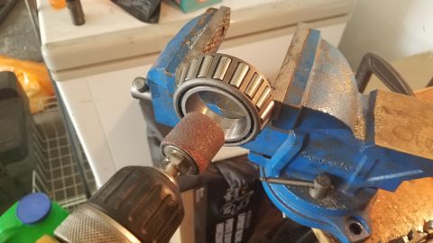 1985 Ford Bronco - A bearing is held in a blue vise while a sanding drum attached to a drill is used to sand the inside of the bearing.