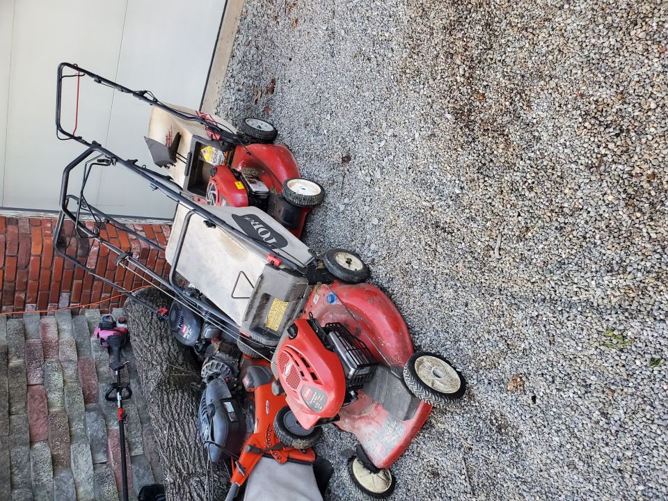 Several red and silver lawnmowers and a string trimmer piled on gravel next to a brick wall