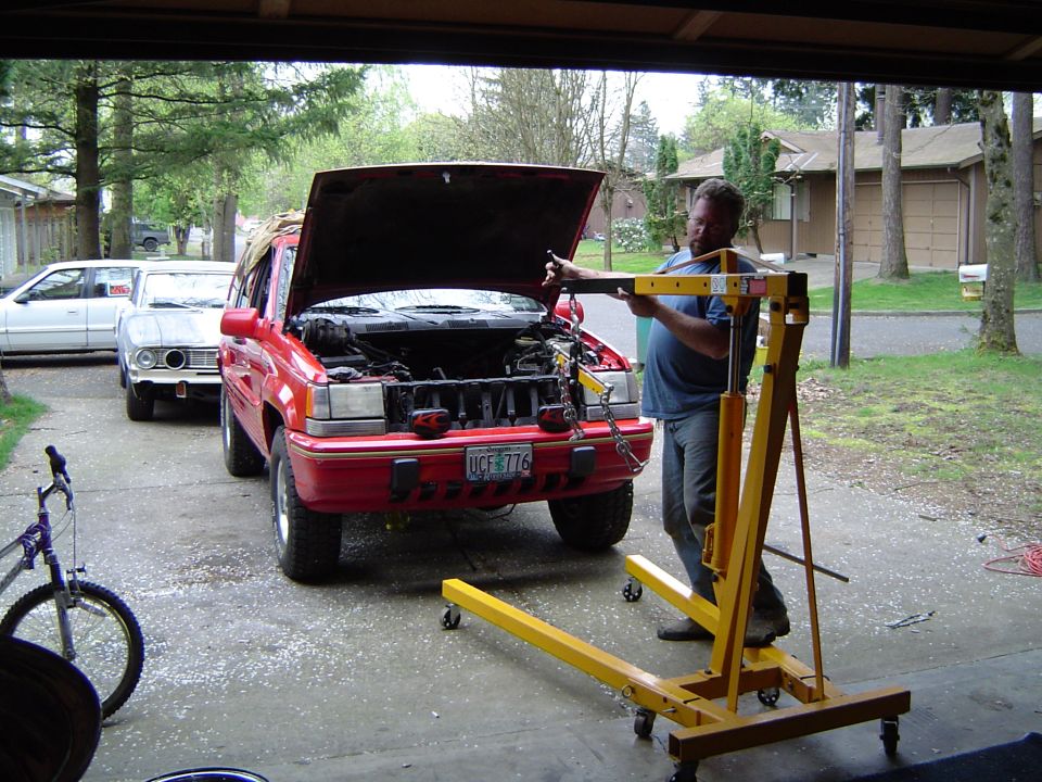 Red SUV with hood up and engine hoist in a driveway, man working, garage behind