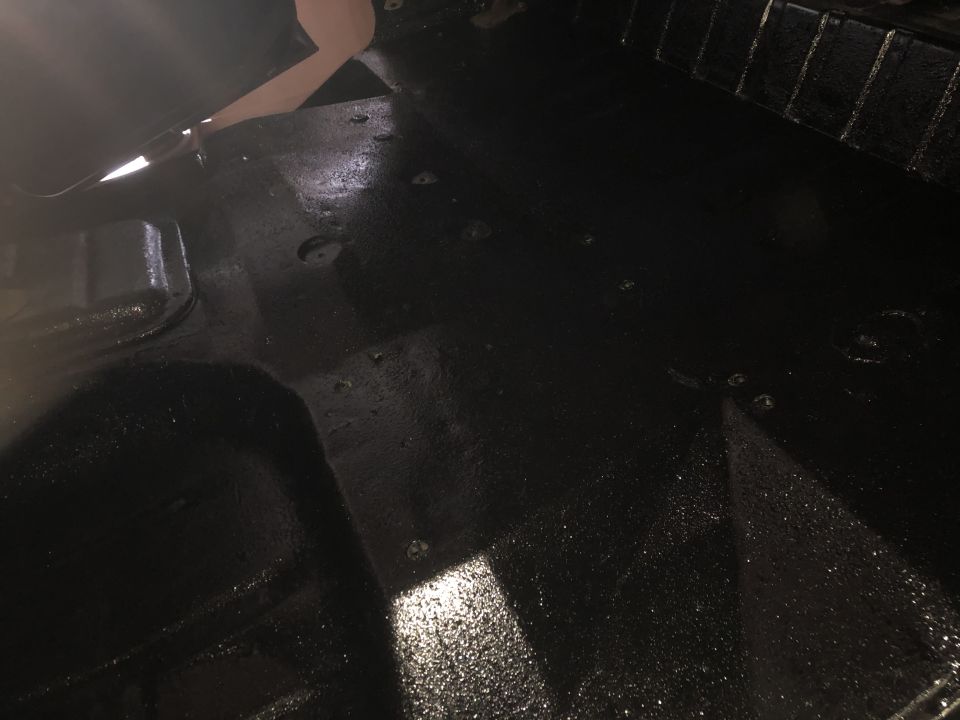 Black painted floor pan with some debris and reflections. Part of a seat frame is visible