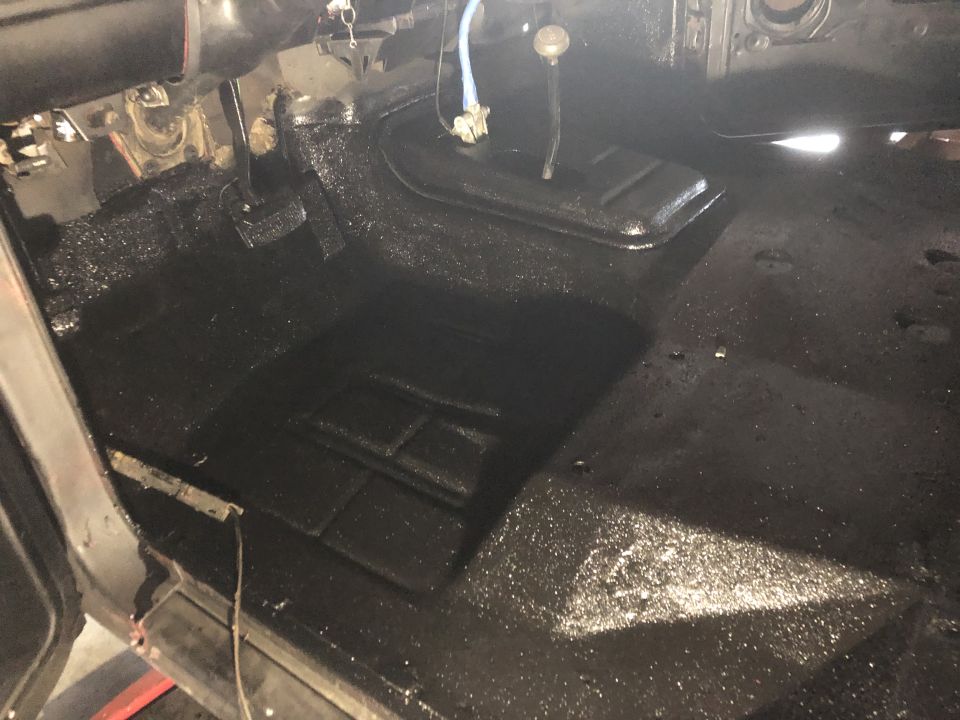 Vehicle floor with black spray-on bedliner applied, showing transmission tunnel and pedals