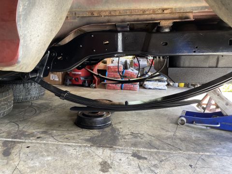 1994 Ford Bronco - The underside of a vehicle shows a black painted frame and leaf spring suspension system, with a brake drum resting on the concrete floor b…