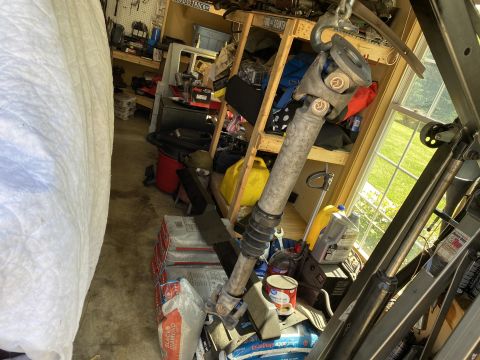 1994 Ford Bronco - A driveshaft with a U-joint is suspended by a chain hoist in a garage setting. Various tools, containers, and bags of material are visible…