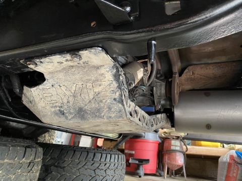 1994 Ford Bronco - The underside of a vehicle shows a dirty skid plate with the "EXO" logo embossed on it, a transfer case, and part of a silver exhaust syste…