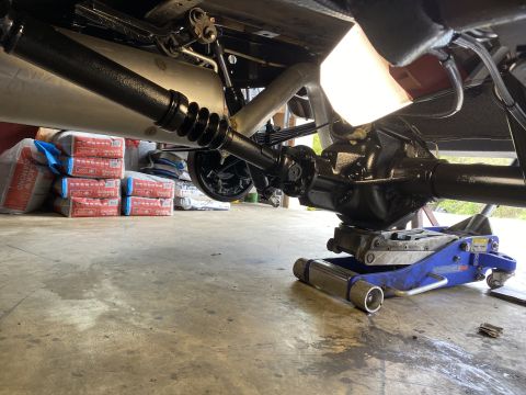 1994 Ford Bronco - The underside of a vehicle, likely a 1994 Ford Bronco, showing the rear differential and driveshaft supported by a blue hydraulic jack. Sev…