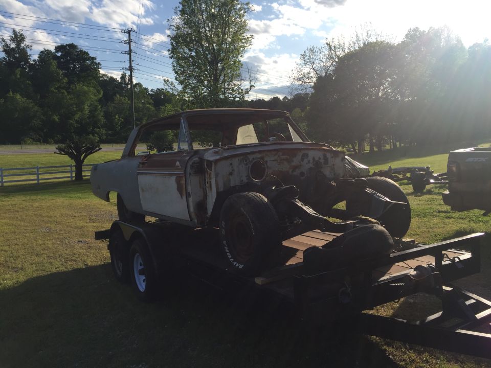 Rusty 1960s coupe body on a trailer, outdoors.