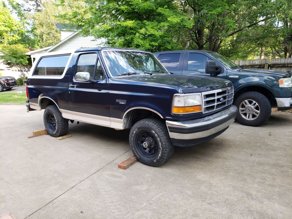 Dark blue Ford Bronco SUV with tan trim on black wheels in a driveway, parked next to a green F-150