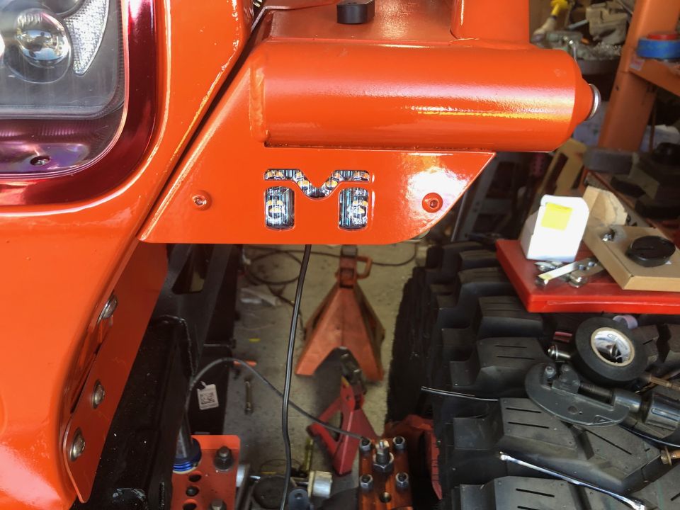 Orange Jeep Wrangler fender with custom "M" LED lights, showing automotive parts and tools in the background.