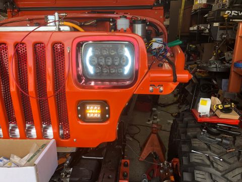1992 Jeep Wrangler - Front view of an orange Jeep Wrangler with custom LED headlights and fog lights. The vehicle is in a workshop setting with various tools an…