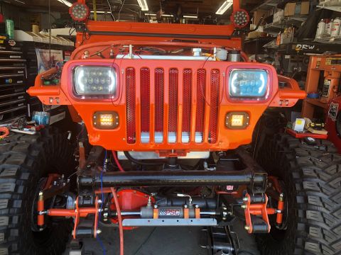 1992 Jeep Wrangler - The front of an orange Jeep Wrangler is shown in a garage setting. It features aftermarket LED headlights, a red grille, and large off-road…