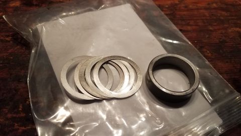 1985 Ford Bronco - A stack of thin metal washers and a single metal spacer are shown inside a clear plastic bag. The items are placed on a wooden surface.