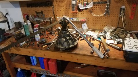 1985 Ford Bronco - A workbench in a garage is cluttered with automotive tools and parts, including a differential and a torque wrench. Various cans and contai…