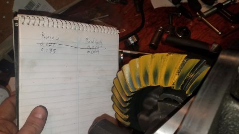 1985 Ford Bronco - A notebook with handwritten measurements for pinion depth and backlash is held up next to a differential gear with yellow and green contact…