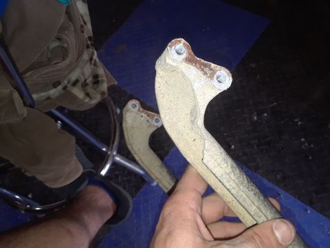 1994 Ford Bronco - A hand holds a worn, tan-colored metal part with two bolt holes at the top, showing signs of rust and damage. Another similar part is visib…
