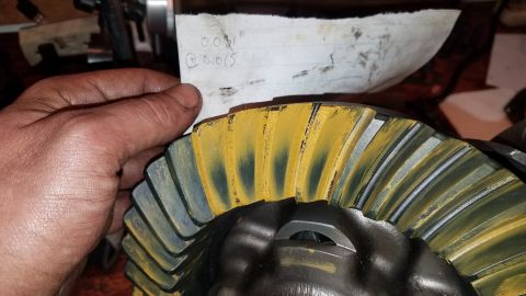 1985 Ford Bronco - A hand holds a piece of paper with handwritten notes over a differential gear. The gear has yellow marking compound applied to its teeth, i…