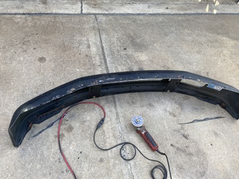 1994 Ford Bronco - A black bumper, likely from a 1994 Ford Bronco, lies on a concrete surface next to a red and black power cord and an angle grinder.