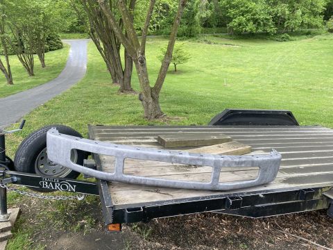 1994 Ford Bronco - A silver aftermarket bumper is laid across a flatbed trailer. The trailer is parked on a gravel and grass area next to a paved driveway.