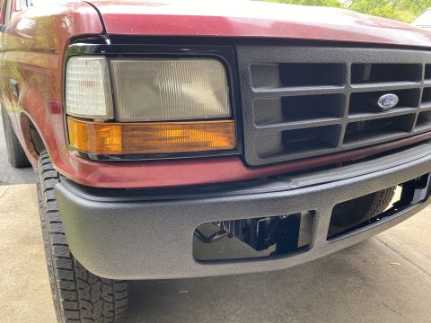 1994 Ford Bronco - Close-up front view of a red Ford Bronco's grille and headlight assembly. The vehicle has a black textured grille with a Ford emblem and a…