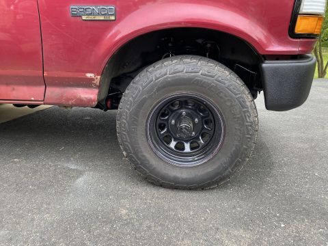 1994 Ford Bronco - Close-up of the front passenger side wheel and fender flare of a red Ford Bronco. The tire is an all-terrain tire on a black steel wheel, a…