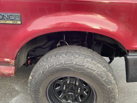 1994 Ford Bronco - Close-up view of the front passenger-side wheel well of a red Ford Bronco. The image shows the tire, suspension components, and fender flar…