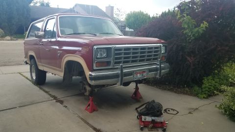 1985 Ford Bronco - A maroon and tan 1980s Ford Bronco is parked on a driveway, supported by jack stands with the front wheels removed. A differential is visib…