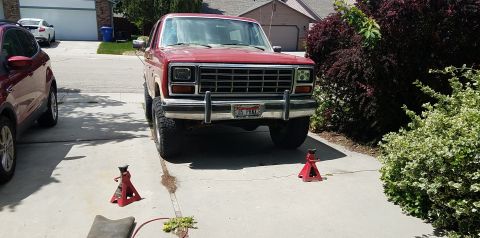 1985 Ford Bronco - A red 1985 Ford Bronco is parked in a driveway with its front end raised on jack stands. Another red SUV is parked to the left of the Bronc…