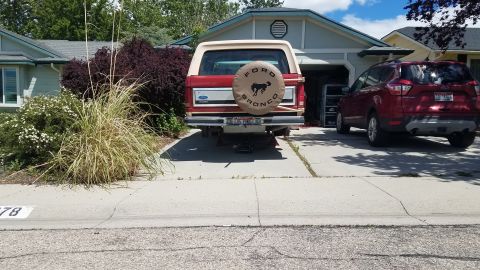 1985 Ford Bronco - The rear of a red 1985 Ford Bronco is shown parked in a driveway with its rear end elevated on jack stands. A red Ford Escape is parked to…