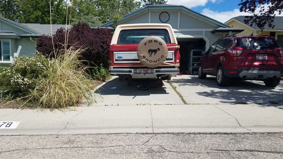 Rear view of a red 1985 Ford Bronco on jack stands, with a red Ford Escape parked nearby.