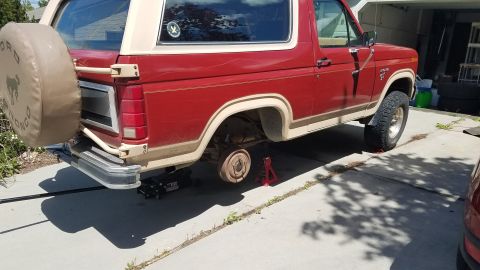 1985 Ford Bronco - The rear passenger side of a red Ford Bronco is shown with the rear wheel removed and the vehicle supported by a jack and jack stands. A sp…