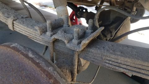1985 Ford Bronco - A close-up view of the rusted leaf spring suspension system on the rear axle of a vehicle, with a differential housing visible in the backg…