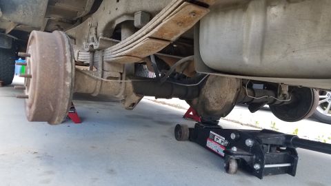 1985 Ford Bronco - The underside of a vehicle is shown, supported by jack stands and a hydraulic jack. A leaf spring suspension, rear axle, drum brake, and fu…
