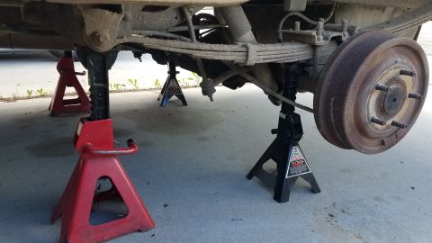 1985 Ford Bronco - The underside of a vehicle is supported by four jack stands on a concrete surface. A rusty drum brake and leaf springs are visible.