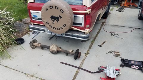 1985 Ford Bronco - A rear axle assembly is laid out on a concrete driveway next to a red Ford Bronco. Tools and a floor jack are scattered around the vehicle.