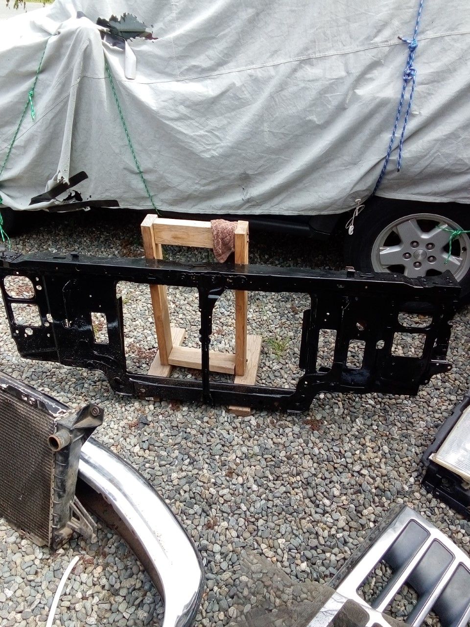 Black radiator support on gravel with vehicle parts and tarp-covered vehicle