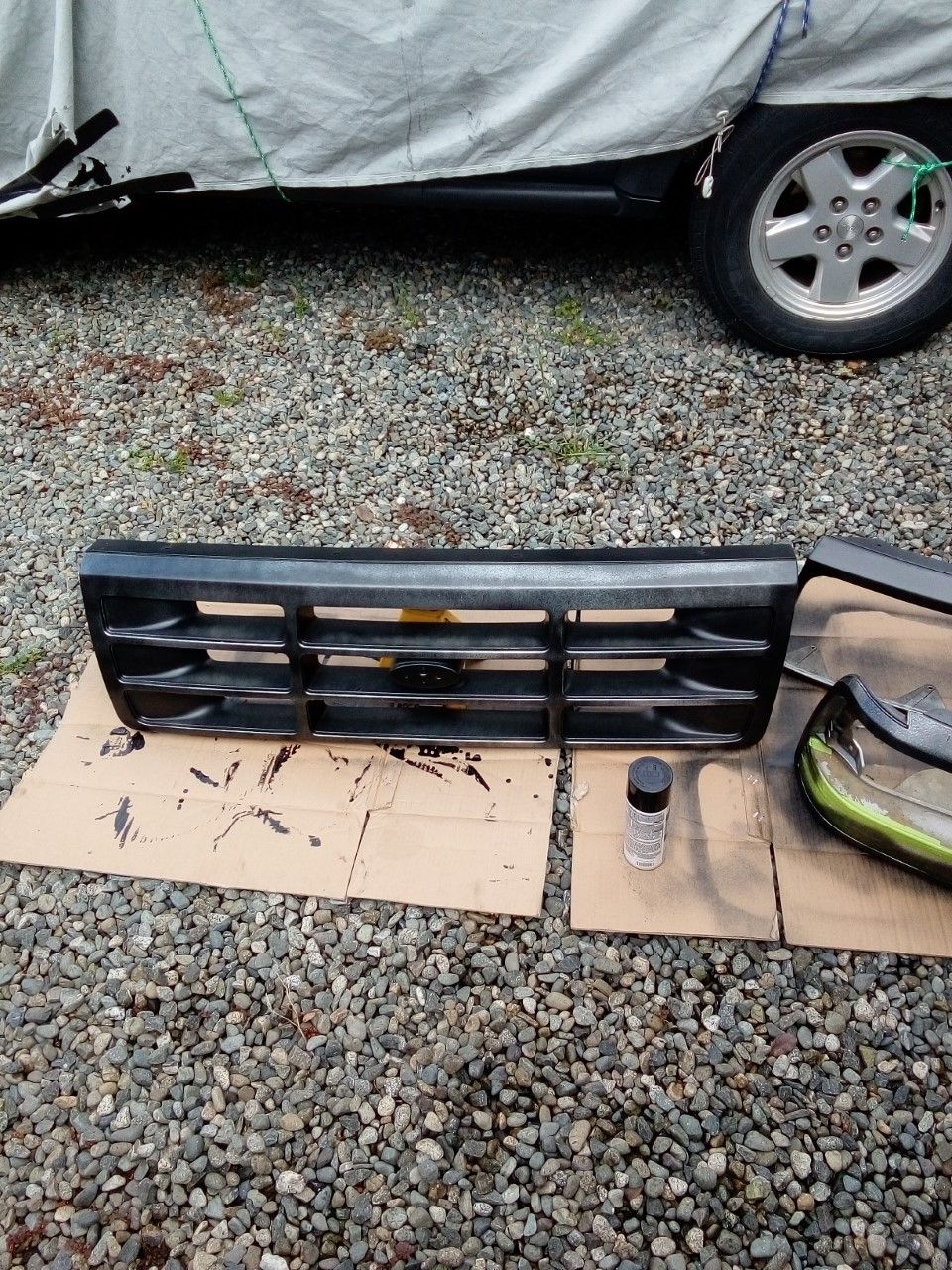 Black Ford Bronco grille on cardboard with spray paint can, vehicle covered by tarp in background.