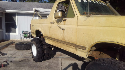 1988 Ford Bronco - A lifted, tan 1988 Ford Bronco with oversized mud tires is parked on a paved driveway next to a house. A spare tire and a hose reel are vis…