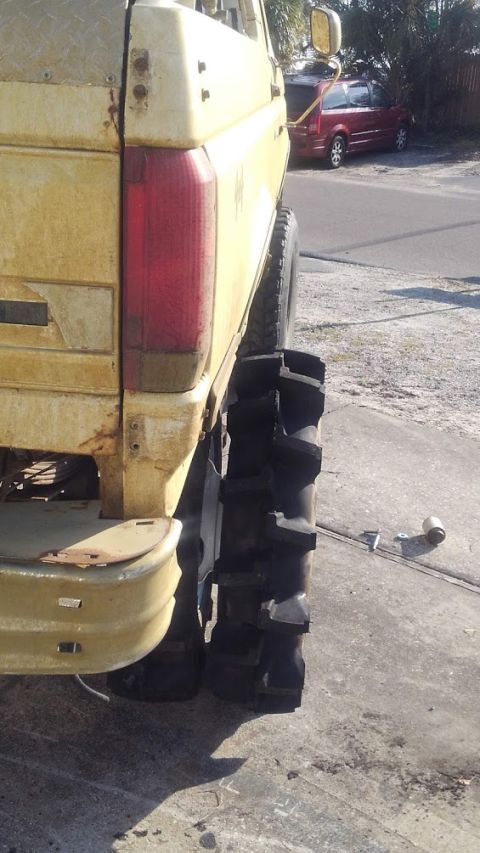 1988 Ford Bronco - The rear passenger side of a weathered yellow pickup truck with oversized, aggressive off-road tires is shown. A red minivan is parked in t…