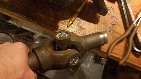 1985 Ford Bronco - A close-up shot shows a mechanic's hand holding a u-joint, likely for a driveshaft. Tools and retaining rings are visible on a wooden workb…