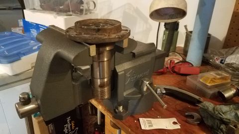 1985 Ford Bronco - A metal part, possibly a hub or bearing assembly, is secured in a black vise on a wooden workbench. Various tools and containers are visibl…