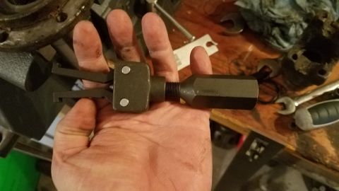 1985 Ford Bronco - A hand holds a dark-colored tool with two prongs and a hexagonal handle, likely a bearing or seal puller, against a wooden workbench with v…