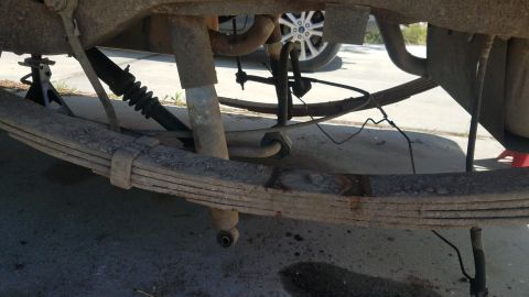 1985 Ford Bronco - The underside of a vehicle shows rusted leaf springs and shock absorbers, with a jack stand supporting the frame. A portion of a tire with…