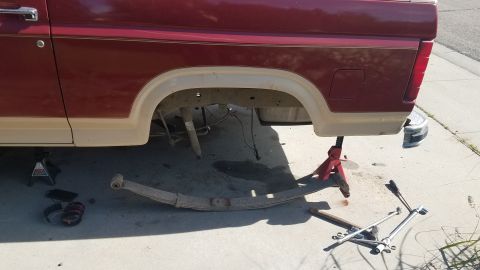 1985 Ford Bronco - The rear passenger side of a maroon and tan pickup truck is jacked up with a leaf spring removed and lying on the ground. Tools and safety…