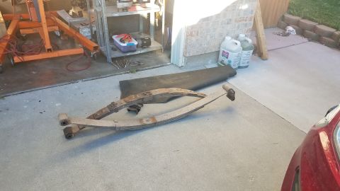 1985 Ford Bronco - Two rusty leaf springs are laid out on a concrete driveway in front of a garage. An orange engine hoist is visible in the background.
