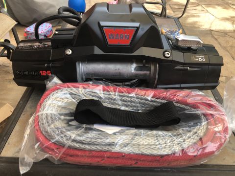 1992 Jeep Wrangler - A black Warn Zeon 12S winch with a red and grey rope is displayed on a table outdoors. The winch has a red WARN logo and a silver freespool…