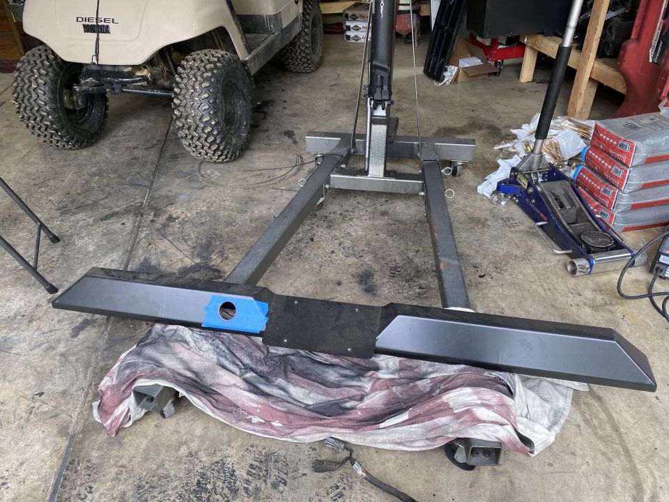 Aftermarket rear bumper on a cloth, engine hoist behind it. Golf cart in background