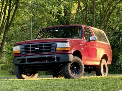 1994 Ford Bronco - This red 1992 Ford Bronco SUV is shown from a three-quarter front angle, parked on a grassy area with trees behind it.