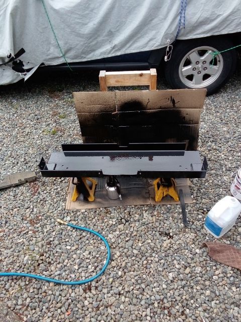 1994 Ford Bronco - A metal component is being spray-painted black while supported by jack stands, with a covered vehicle nearby.