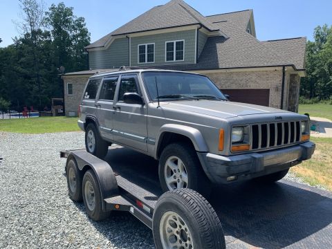 2000 Jeep Cherokee - A silver 2000 Jeep Cherokee XJ is loaded onto a trailer, parked in front of a residential building.